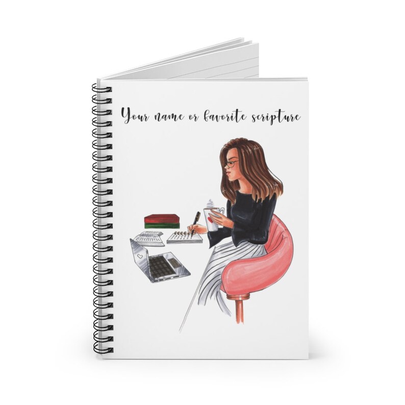 May include: A white spiral bound notebook with a colorful illustration of a woman sitting at a desk with a laptop, a cup of coffee, and a pen. The text "Your name or favorite scripture" is at the top of the notebook.