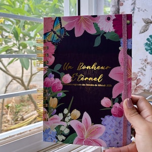 Peut inclure: Un journal à spirales avec une couverture ornée de fleurs et l'inscription "Un Bonheur Éternel". Le design floral comprend des lys roses, des hortensias bleus et un papillon. La reliure est dorée.