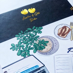 May include: A spiral-bound notebook with a black cover featuring gold text and apple illustrations. The notebook is open, revealing a detailed illustration of a laptop, a plant, a plate with food, and a fork. The text on the cover reads "Apples & Gold Studio."