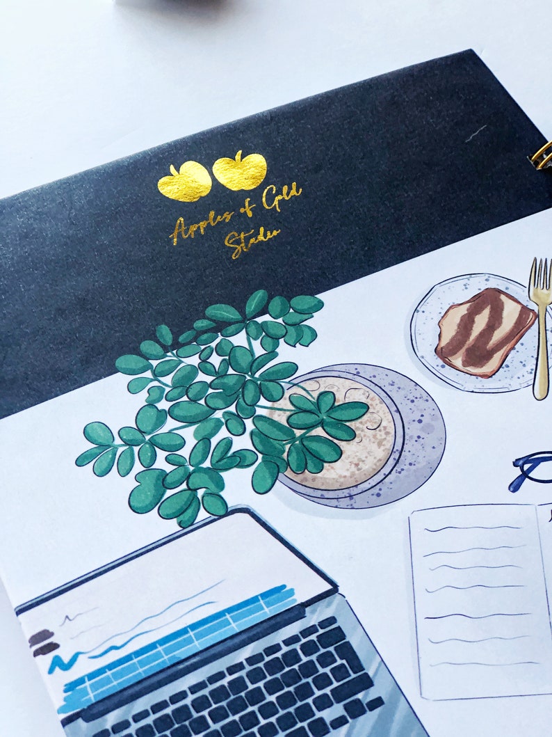 May include: A spiral-bound notebook with a black cover featuring gold text and apple illustrations. The notebook is open, revealing a detailed illustration of a laptop, a plant, a plate with food, and a fork. The text on the cover reads "Apples & Gold Studio."
