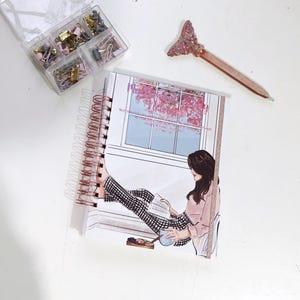 May include: A spiral-bound notebook with a woman reading by a window illustration. A clear plastic container holds paper clips and binder clips. A pen with a glittery tail is also present.