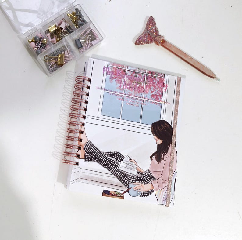 May include: A spiral-bound notebook with a woman reading by a window illustration. A clear plastic container holds paper clips and binder clips. A pen with a glittery tail is also present.