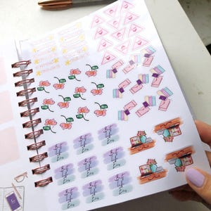 May include: A planner with various stickers, including floral, money, and text designs. The stickers feature phrases like "Best Life Ever" and "To Remind". A pen rests on the planner's page.