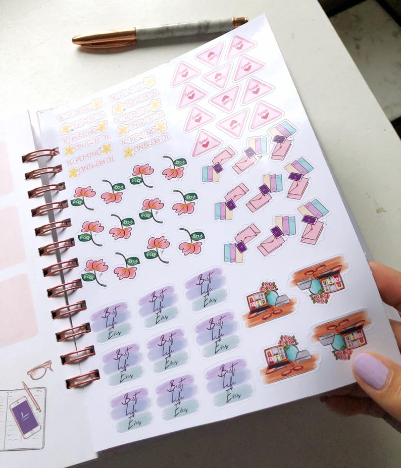 May include: A planner with various stickers, including floral, money, and text designs. The stickers feature phrases like "Best Life Ever" and "To Remind". A pen rests on the planner's page.