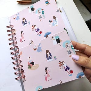 May include: A spiral-bound notebook with a pink cover featuring illustrations of women in various poses. The notebook has a rose gold spiral binding and a pen resting on top. The pocketed cover is open, revealing the interior pages.