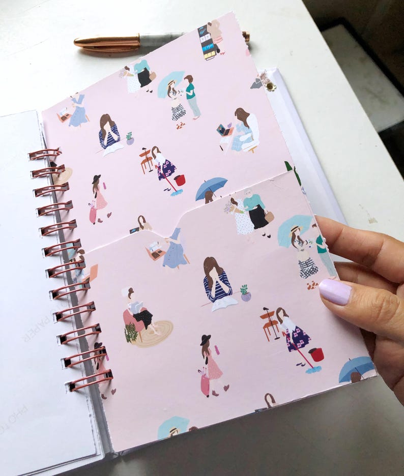 May include: A spiral-bound notebook with a pink cover featuring illustrations of women in various poses. The notebook has a rose gold spiral binding and a pen resting on top. The pocketed cover is open, revealing the interior pages.