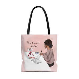 Pu&ograve; includere: Una borsa tote rosa con un manico nero presenta un'illustrazione di una donna che scrive su un diario a un tavolo. La donna ha i capelli castani ricci e indossa un maglione marrone chiaro. Il testo "Your favorite scripture" &egrave; stampato sulla borsa.