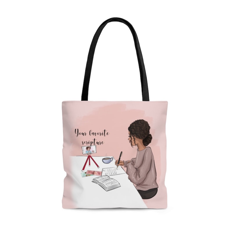 Pu&ograve; includere: Una borsa tote rosa con un manico nero e un design di una donna che scrive su un quaderno. Il design include anche un telefono su un treppiede, una tazza di caff&egrave; e il testo "Your favorite scripture".