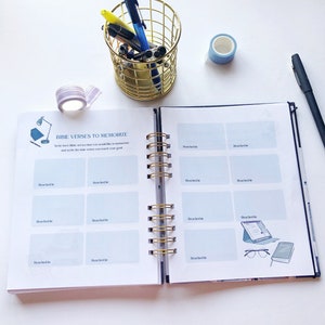 May include: Open spiral-bound notebook with the text "BIBLE VERSES TO MEMORIZE" and fill-in-the-blank sections. A gold wire pen holder, pens, and washi tape are on the white surface. A black pen rests on the right side.