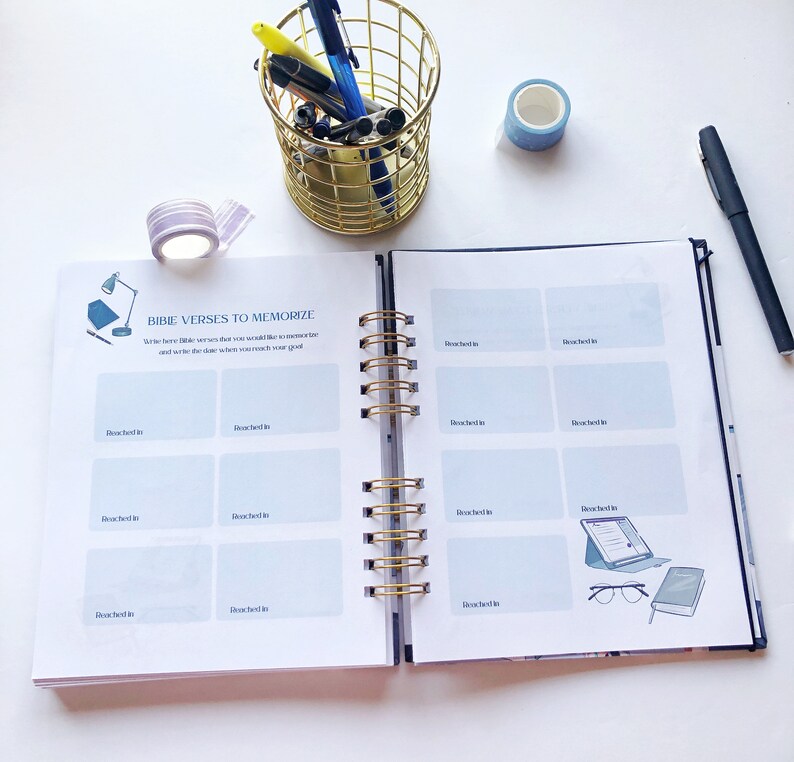May include: Open spiral-bound notebook with the text "BIBLE VERSES TO MEMORIZE" and fill-in-the-blank sections. A gold wire pen holder, pens, and washi tape are on the white surface. A black pen rests on the right side.
