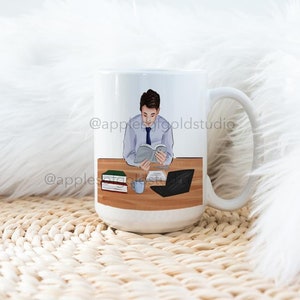 May include: White ceramic mug featuring an illustration of a person reading at a desk with a laptop, books, and a coffee cup. The mug has a large handle and a glossy finish. The illustration includes the text "@applegoldstudio".
