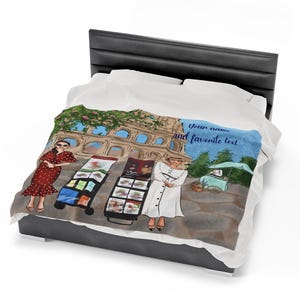 May include: A white and blue blanket with a colorful illustration of a street scene in Rome, Italy. The illustration features two women, one in a red and white polka dot dress and the other in a white dress, selling goods from carts. The Colosseum is in the background. The text "Your name and favorite text" is printed on the blanket.