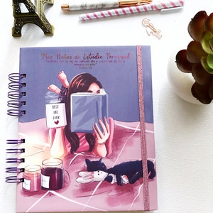 May include: A spiral-bound notebook with a purple cover featuring an illustration of a person reading in bed. The text "Mis Notas de Estudio Personal" is at the top. The notebook is next to pens, candles, and a decorative Eiffel Tower.