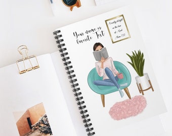 May include: Spiral-bound notebook with a printed illustration of a person reading in a teal chair. The notebook has the text "Your name or favorite Text" and a gold framed quote. A gold binder clip and sunglasses are also visible.