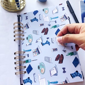 May include: A spiral-bound notebook with a light blue cover featuring a pattern of office-related illustrations, including laptops, coffee cups, and bow ties. A gold pen holder and blue washi tape are visible.