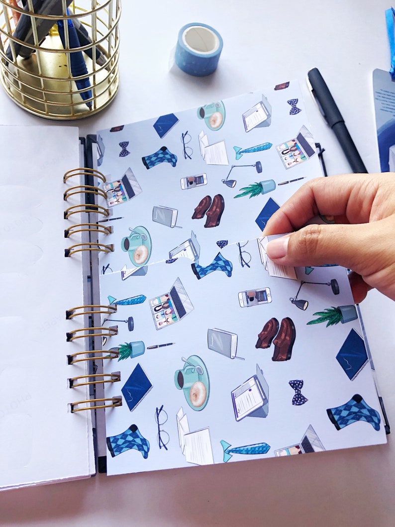 May include: A spiral-bound notebook with a light blue cover featuring a pattern of office-related illustrations, including laptops, coffee cups, and bow ties. A gold pen holder and blue washi tape are visible.