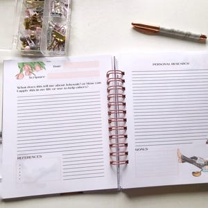 May include: Open spiral-bound journal with lined pages, featuring sections for scripture, personal research, and goals. A rose gold pen and a container of paper clips and push pins are on the white surface. The journal is open to a double-page spread.