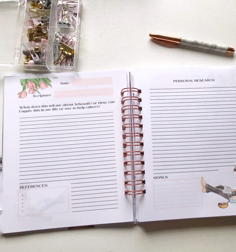 May include: Open spiral-bound journal with lined pages, featuring sections for scripture, personal research, and goals. A rose gold pen and a container of paper clips and push pins are on the white surface. The journal is open to a double-page spread.