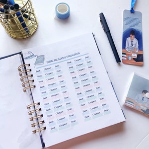 May include: An open spiral-bound planner with a "BIBLE READING PROGRAM" layout, featuring columns for "Book," "Chapter," and "Date." A blue and white patterned washi tape roll, a black pen, and a bookmark with a reading illustration are also visible.