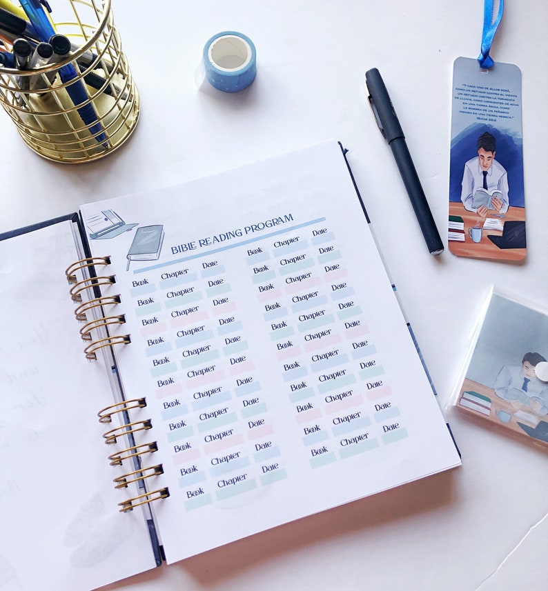 May include: An open spiral-bound planner with a "BIBLE READING PROGRAM" layout, featuring columns for "Book," "Chapter," and "Date." A blue and white patterned washi tape roll, a black pen, and a bookmark with a reading illustration are also visible.