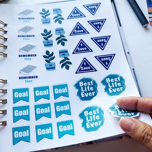 May include: A collection of planner stickers with various designs and text. The stickers include blue triangles with "Save", blue flags with "Goal", and stickers with "Best Life Ever". There are also stickers with "Remember This" and "To Remind".