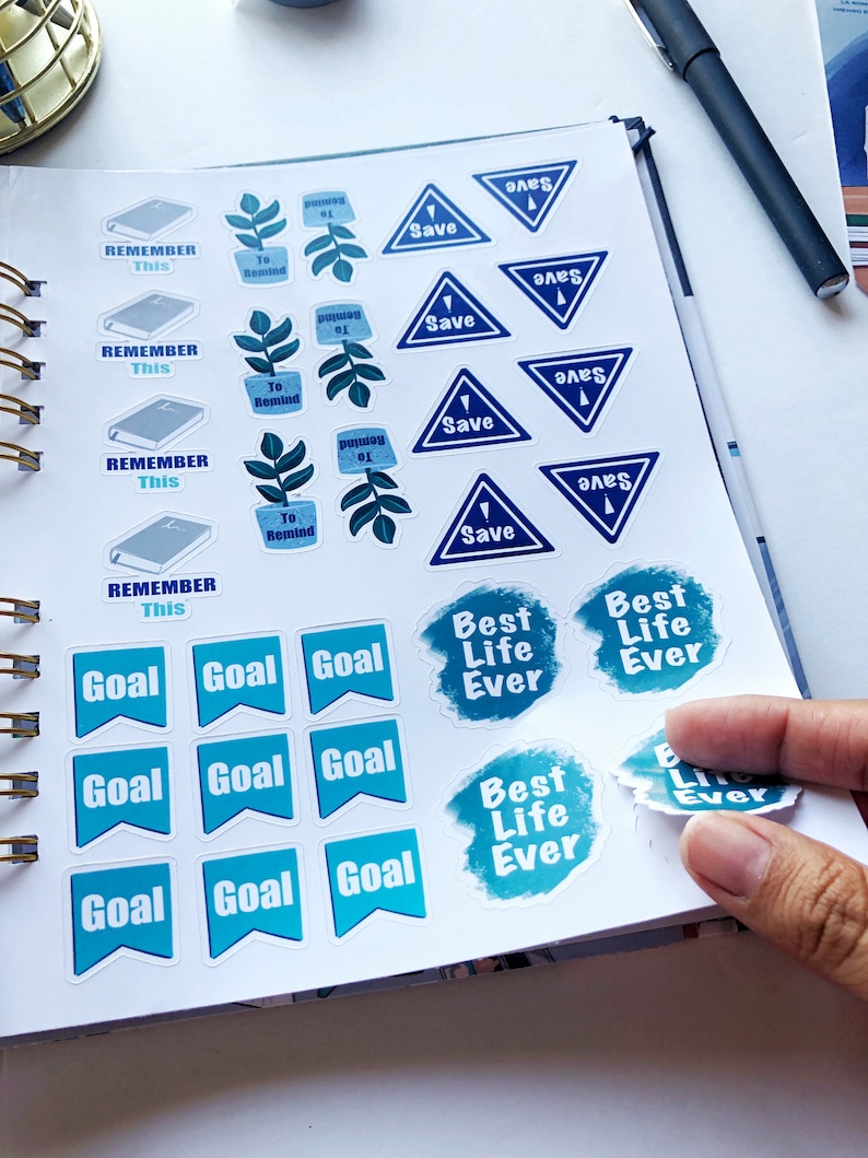 May include: A collection of planner stickers with various designs and text. The stickers include blue triangles with "Save", blue flags with "Goal", and stickers with "Best Life Ever". There are also stickers with "Remember This" and "To Remind".