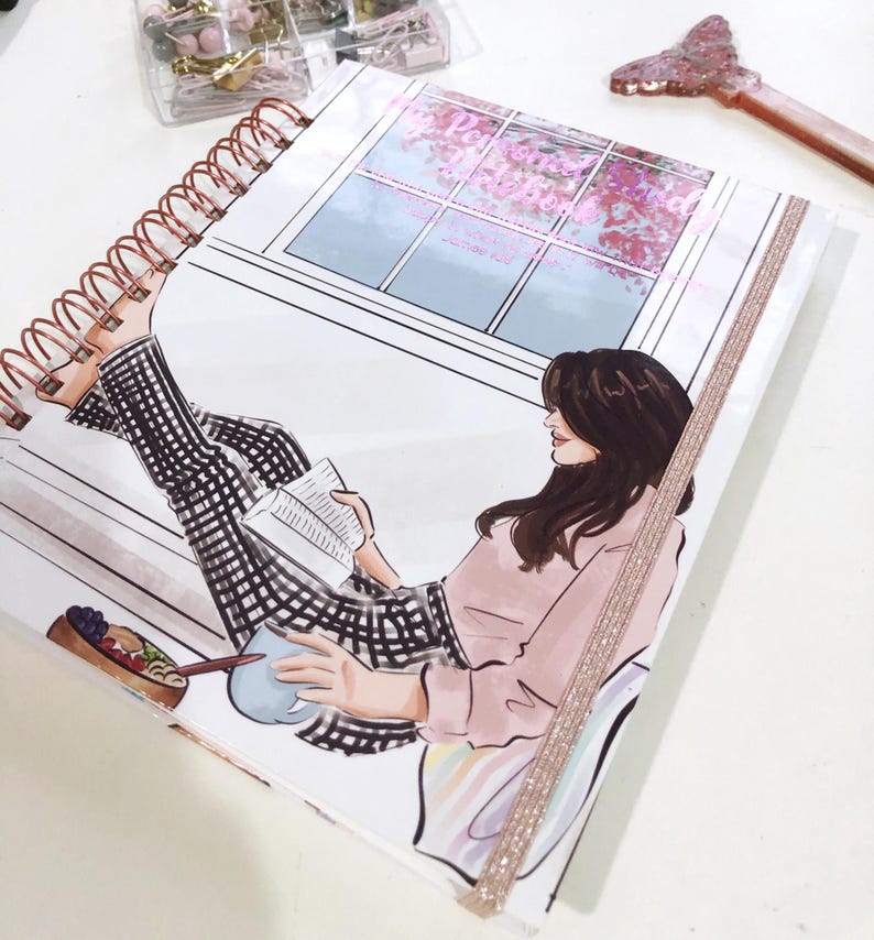 May include: A spiral-bound notebook featuring an illustration of a person reading. The illustration shows a person in a pink top and black and white checkered pants, reading a book near a window with pink flowers. A decorative butterfly-shaped item is visible.