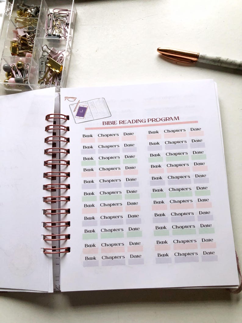 May include: Open spiral-bound notebook with a Bible reading program layout. The pages are white with a grid of boxes for tracking book, chapters, and date. A clear container of paperclips and a pen are in the background.