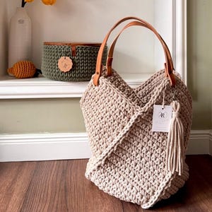 May include: A beige crochet tote bag featuring a textured knit pattern, two tan leather handles, and a matching beige tassel hanging from a silver clasp. A white rectangular tag with the letter R is attached to the side of the bag.