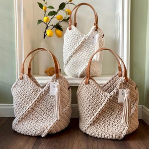 May include: Three crochet tote bags with tan leather handles and matching tassels. One bag is white, and two are beige. Each bag features a textured, thick yarn construction with a diagonal overlapping front panel design and a small branded tag.