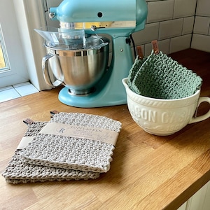 Könnte beinhalten: Ein blauer Küchenmixer mit einer silbernen Schüssel und einem durchsichtigen Spritzschutz aus Kunststoff. Im Vordergrund befinden sich zwei beigefarbene gehäkelte Topflappen mit Lederriemen und einem braunen Etikett mit der Aufschrift "Die Topplappen Handmade Set 2 Stück". Im Hintergrund steht eine weiße Keramikschüssel mit einem grünen gehäkelten Topflappen darin.