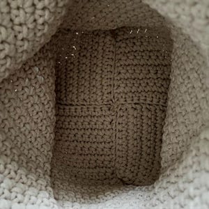 May include: A beige crochet bag interior showing the bottom base and side walls. The bag is constructed with a dense, uniform stitch pattern in a light tan yarn, featuring a square-shaped base and flexible, soft-textured fabric walls.