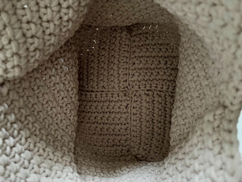 May include: A beige crochet bag interior showing the bottom base and side walls. The bag is constructed with a dense, uniform stitch pattern in a light tan yarn, featuring a square-shaped base and flexible, soft-textured fabric walls.