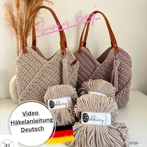 Könnte beinhalten: Zwei beige gehäkelte Tragetaschen mit braunen Ledergriffen und Quasten. Die Taschen werden mit mehreren Spulen beigefarbenem Bobbiny Premium Makramee-Garn präsentiert. Das Bild enthält auch den Text "Sunny Bag" und "Video Häkelanleitung Deutsch".