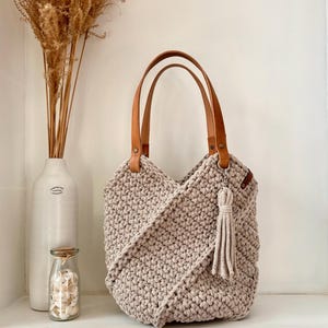 May include: A beige crochet tote bag featuring a textured, knotted pattern with a diagonal overlapping front design. The bag includes two tan leather shoulder straps attached with metal rivets and a matching beige crochet tassel hanging from a ring.