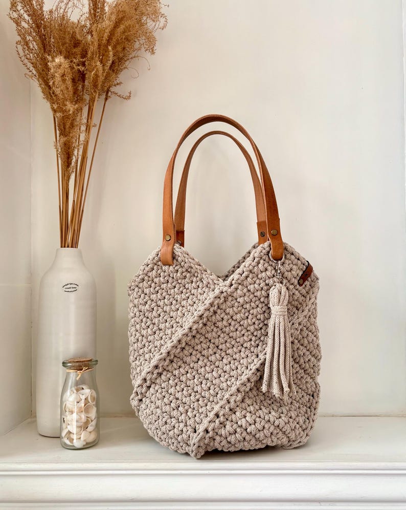 May include: A beige crochet tote bag featuring a textured, knotted pattern with a diagonal overlapping front design. The bag includes two tan leather shoulder straps attached with metal rivets and a matching beige crochet tassel hanging from a ring.