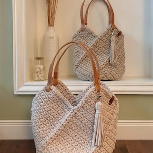 K&ouml;nnte beinhalten: Zwei beige geh&auml;kelte Tragetaschen mit strukturiertem Stichmuster, hellbraunen Ledergriffen und einer passenden beigen Quaste an einem silbernen Metallverschluss. Die Taschen haben ein V-f&ouml;rmiges Frontdesign und bestehen aus dickem Kordelmaterial.
