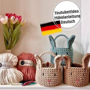 Könnte beinhalten: Drei gehäkelte Hasen-Körbe in Beige, Hellblau und Braun, mit rosa Nasen und Henkeln. Zwei Knäuel Garn in Weiß und Koralle sowie ein Strauß rosa und weißer Tulpen sind ebenfalls zu sehen. Der Text im Bild lautet "YoutubeVideo Häkelanleitung Deutsch".