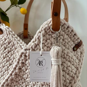May include: A beige crocheted tote bag featuring tan leather handles secured with metal rivets. A matching crocheted tassel hangs from a silver clasp. A white rectangular tag displays the logo WR and the text We and With Rose DESIGN HANDMADE BAGS.