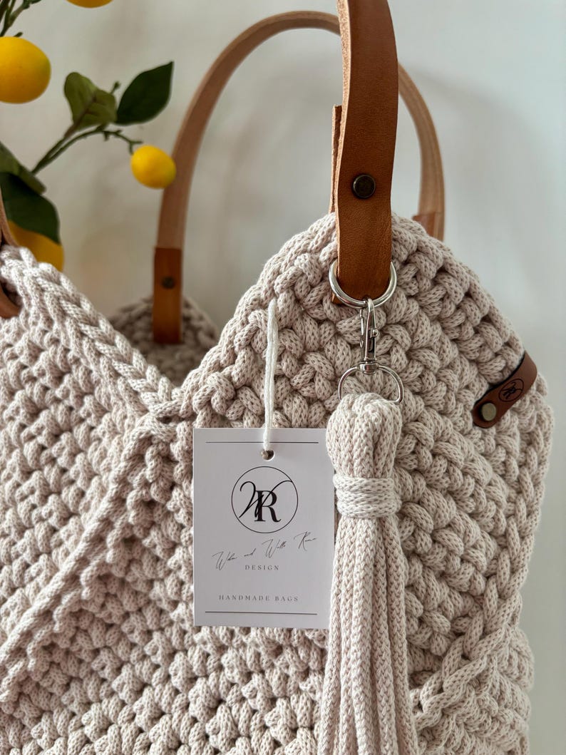 May include: A beige crocheted tote bag featuring tan leather handles secured with metal rivets. A matching crocheted tassel hangs from a silver clasp. A white rectangular tag displays the logo WR and the text We and With Rose DESIGN HANDMADE BAGS.