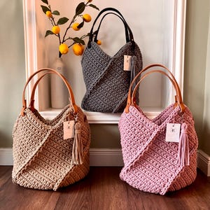 May include: Three crochet tote bags in beige, dark gray, and dusty pink. Each bag features a thick textured knit pattern, two tan or black leather-like handles, and a matching hanging tassel with a small white rectangular brand tag attached.