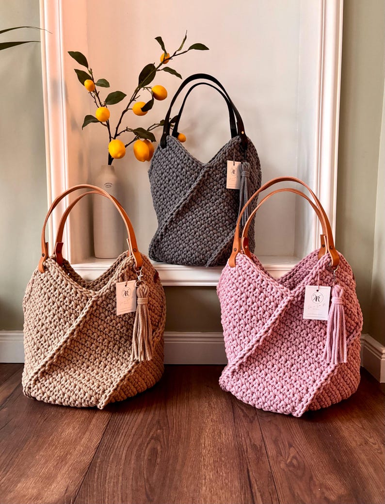 May include: Three crochet tote bags in beige, dark gray, and dusty pink. Each bag features a thick textured knit pattern, two tan or black leather-like handles, and a matching hanging tassel with a small white rectangular brand tag attached.