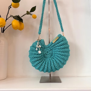 May include: A teal crochet handbag with a shell-like spiral design and a single braided shoulder strap. The bag features a gold-toned metal tag labeled W&W Collection and a dangling charm accessory with white faux pearls and a small circular metal pendant.