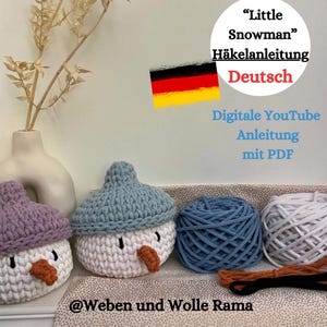 May include: Two crocheted snowman ornaments with blue and purple hats, white yarn, and orange noses. The image also includes balls of yarn in blue and white, and a brown and black cord. The text in the image reads "Little Snowman" Häkelanleitung Deutsch Digitale YouTube Anleitung mit PDF @Weben und Wolle Rama.
