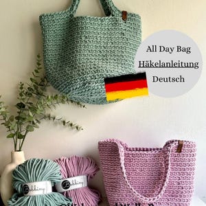 Peut inclure: Deux sacs cabas en crochet, l'un vert et l'autre rose, avec des pelotes de laine assorties. Les sacs sont faits avec une corde de coton épaisse et ont des anses en cuir. L'image comprend également le texte "All Day Bag Häkelanleitung Deutsch" et un drapeau allemand.