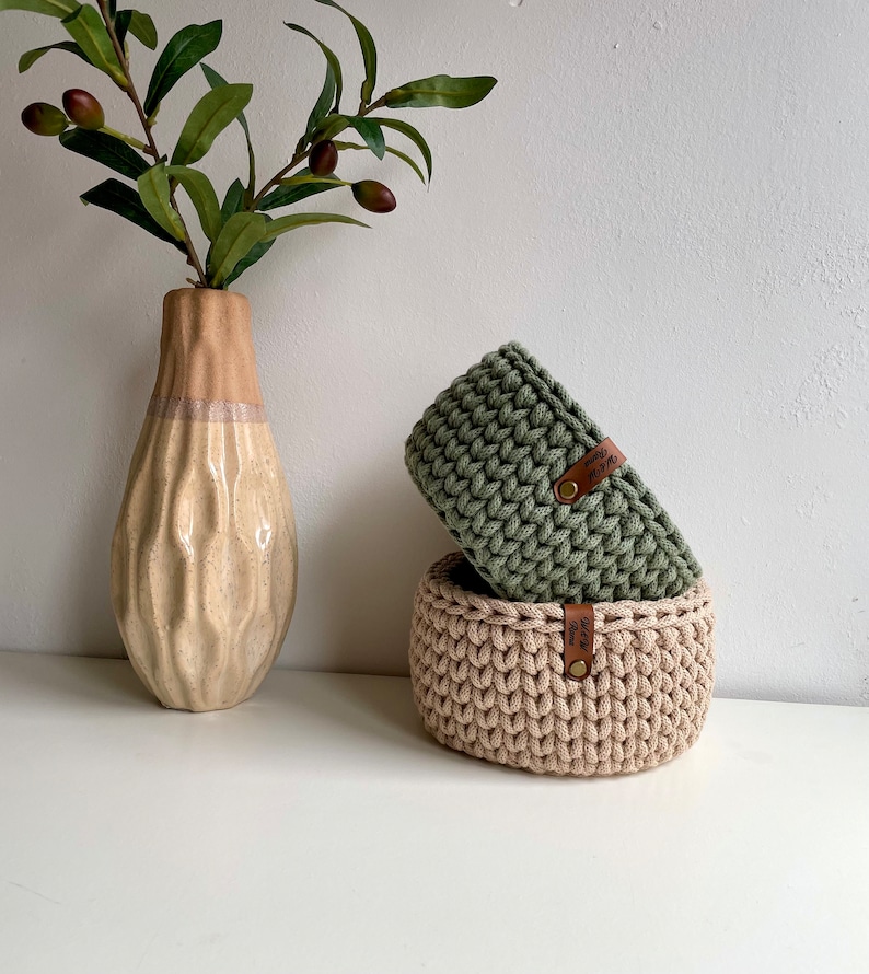 Puede incluir: Dos cestas de crochet, una verde y otra beige, con etiquetas de cuero que dicen "Made with Love". Las cestas est&aacute;n apiladas una encima de la otra sobre una superficie blanca.