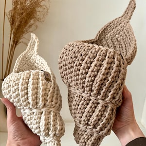 Könnte beinhalten: Zwei handgefertigte Muschelkörbe aus Häkelarbeit in Creme und Beige. Die Körbe haben ein strukturiertes, geripptes Design und eine spitze Spitze. Die Körbe werden von Händen gehalten. Der Hintergrund umfasst getrocknete Pflanzen in einer Vase.
