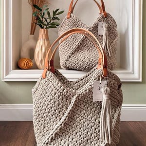 May include: Two beige crochet handbags featuring a textured knit pattern, tan leather handles secured with metal rivets, and a matching beige fabric tassel attached to a white rectangular tag with the letter R printed in black on the front.