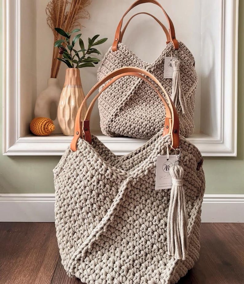 May include: Two beige crochet handbags featuring a textured knit pattern, tan leather handles secured with metal rivets, and a matching beige fabric tassel attached to a white rectangular tag with the letter R printed in black on the front.