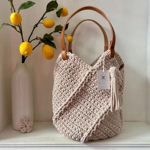 May include: A beige crochet handbag featuring a textured knit pattern, two tan leather handles, and a matching tassel charm. A small white rectangular tag with the letter R is attached to the handle. The bag has a structured, rounded shape.